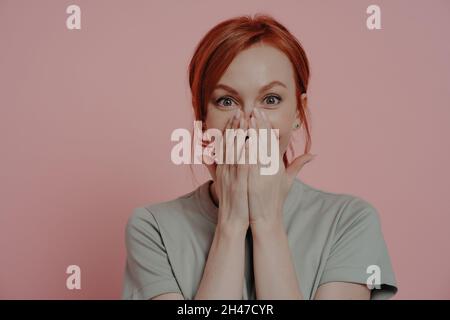 Young pretty ginger woman covering mouth with both hands, being in shock and disbelief Stock Photo