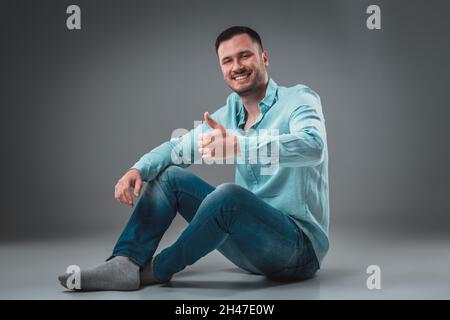 Appealing casual young man sitting on the floor, looking to the camera smiling Stock Photo