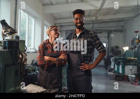 Portrait of young biracial industrial colleagues working indoors in metal workshop, smiling and looking at camera. Stock Photo