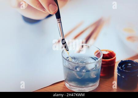 The artist washes the brush in a glass of water, washing off the blue gouache from it. Drawing on a white sheet of paper. Creativity and art. Time for Stock Photo