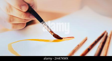 The artist draws lines with a brush in red and yellow watercolor paint on a sheet of white paper illuminated by daylight. Creativity and art. Time for Stock Photo