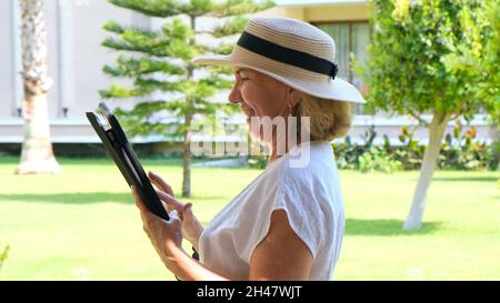 Senior female blonde businesswoman in hat uses tablet pc, digital tablet for business work or study in her own green garden. woman aged 50-55. The Stock Photo