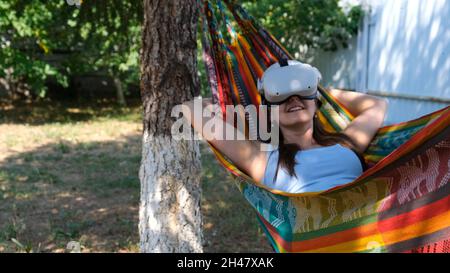 funny woman wearing virtual reality glasses rests in a hammock near a tree, imagining a dream come true, an invented world. The concept of getting Stock Photo