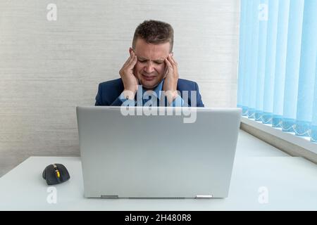 A man in a blue suit with a sore head in the office. The concept of overwork at work. A tired businessman holds his head with his hands. Stock Photo