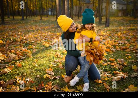 Cheerful loving mother hugs her lovely cute baby girl sitting on her knees while relaxing in the autumn park and gathering a bouquet of falling dry ma Stock Photo
