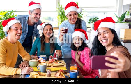 merry christmas! happy multiethnic group of friends wearing xmas santa hat making a selfie portrait sitting on a table at sweet breakfast time Stock Photo