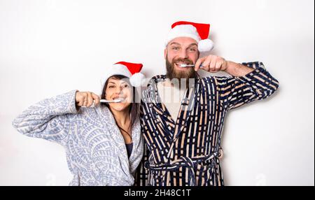 merry christmas! Young adult couple celebrating xmas time wearing santa claus hat standing isolated against background wall brushing teeth. family Stock Photo