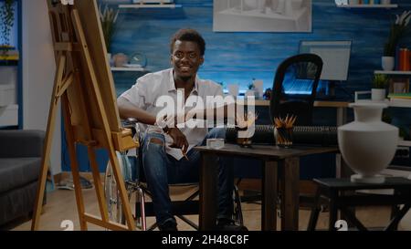 Portrait of disabled african american artist at creativity studio. Black young person with handicap sitting in wheelchair and drawing vase design on canvas for modern masterpiece project Stock Photo