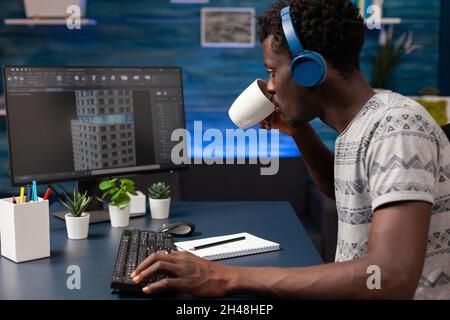 African american engineer drinking coffee analyzing building construction plan working remote from home at architecture sketch late at night. Man architect with headset developing skyscraper model Stock Photo