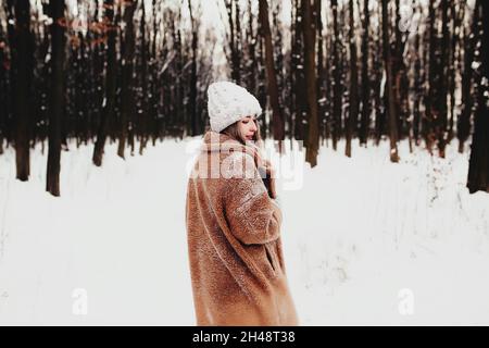 Photo of young beautiful woman in fur coat standing on the white snow in winter forest. Girl is smiling and happy with closed eyes. Female has winter Stock Photo