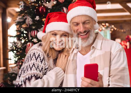 Happy mature couple in santa hats using cell phone near Christmas tree at home. Stock Photo