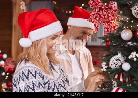 Happy mature old couple in santa hats decorating Christmas tree at home. Stock Photo