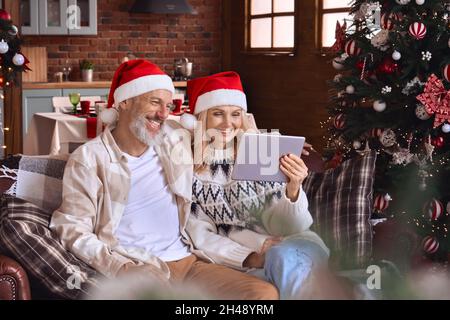 Happy mature old couple using digital tablet relaxing on couch on Christmas. Stock Photo