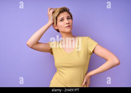 Confused short haired young female touching head having dilemma difficult choice dressed in casual yellow t-shirt isolated over purple studio backgrou Stock Photo