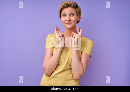 Portrait of glad smiling caucasian woman crosses fingers, believes in good luck and success, looks hopefully straightly at camera, isolated over purpl Stock Photo