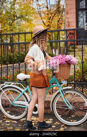 Side view of young woman with curly hair in beige hat and stylish dressed standing near vintage blue bicycle with beautiful bouquet pink flowers. Concept of walking in centre city or park in autumn. Stock Photo