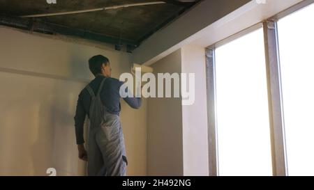 Painter working with paint roller to paint the wall of a room. Do it yourself concept. Man paints the wall with a roller at the window. Stock Photo