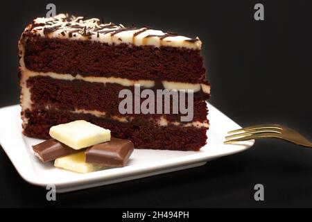 A slice of chocolate cake with cream on a white plate, covered with cream and white chocolate on a black background. festive food concept. Stock Photo