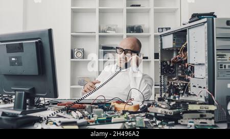 Distracted technician sitting at desk and talking on the phone instead of repairing a broken computer Stock Photo