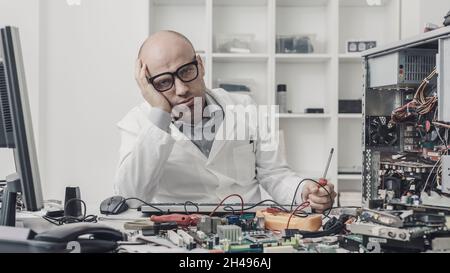 Frustrated exhausted technician trying to repair a computer, his desk is full of computer parts Stock Photo