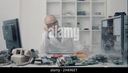Stressed frustrated technician sitting at desk in his office, he has damaged a computer and he is unable to repair it Stock Photo