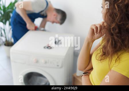 Professional plumber repairing a broken washing machine, the customer is looking at him, repair service concept Stock Photo