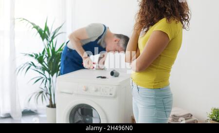 Professional plumber repairing a broken washing machine, the customer is looking at him, repair service concept Stock Photo