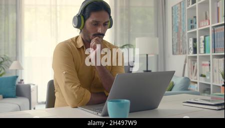 Man sitting at desk at home and connecting with his laptop, he is wearing wireless headphones and watching videos online Stock Photo