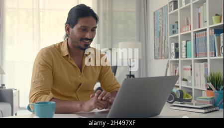 Man sitting at desk at home and video calling his friends online, he is looking at the laptop screen and smiling Stock Photo