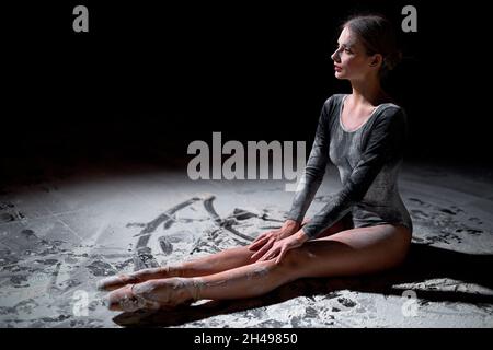 Perfect calm caucasian ballet dancer in black bodysuit in flour in black studio, sitting on floor relaxed, having rest in the end of performance. peop Stock Photo