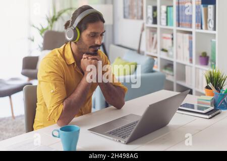 Man sitting at desk at home and connecting with his laptop, he is wearing wireless headphones and watching videos online Stock Photo
