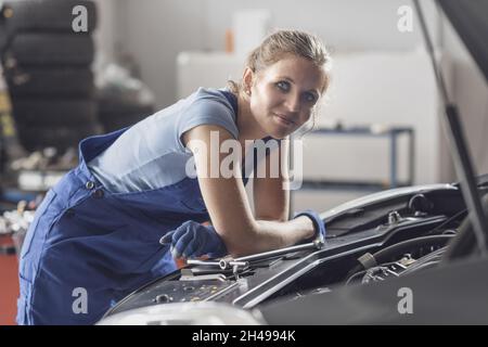 Female mechanic working at the auto repair shop, she is checking a car engine and smiling at camera Stock Photo
