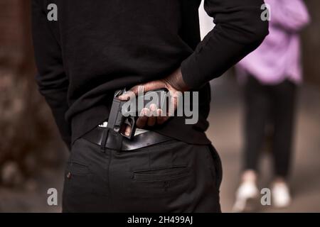 Robbery or criminal man in black hoodie is holding gun standing behind the woman, going to commit a crime. rear view on male in black outfit, close-up Stock Photo