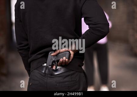 Robbery or criminal man in black hoodie is holding gun standing behind the woman, going to commit a crime. view from back on male in black outfit, clo Stock Photo