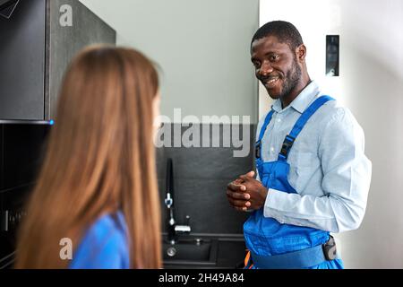 Caucasian Client Woman is grateful to smiling afro plumber for repair glitch in kitchen, confident handyman in blue overalls having talk with female c Stock Photo