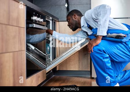 focused african man or service worker in special clothing installs, disassembles or performs maintenance of dishwasher built into kitchen furniture, s Stock Photo