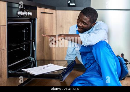 Nice Contractor Handyman In Overalls Looking inside of electric oven examining before repair while writing information about oven, pointing index fing Stock Photo
