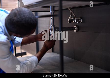 Handsome Hardworking Plumber Installing The Sink In Bathroom Close-up Photo, Cropped Black Man. Guy Is Repairing, Fixing Sink. Skilled Handyman During Stock Photo