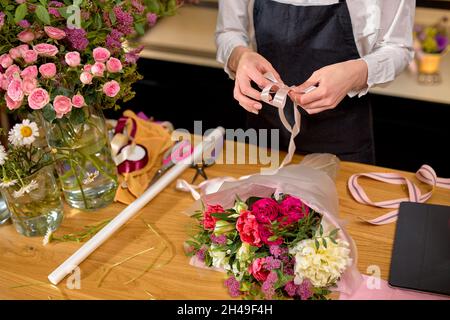 Female florist at work arranging various flowers in bouquet, decorating bouquet. close-up hands of young woman standing behind desk making bouquet of Stock Photo