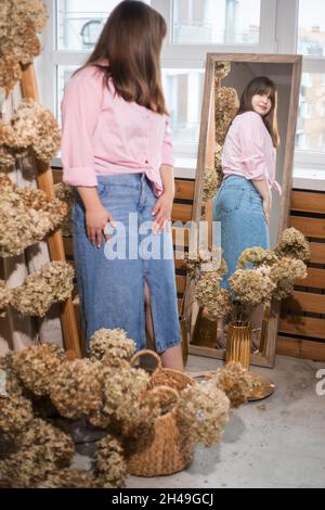 Portrait of young woman decorator florist looks at her reflection in mirror, dressed in blue denim skirt and pink shirt with back view around dried hy Stock Photo