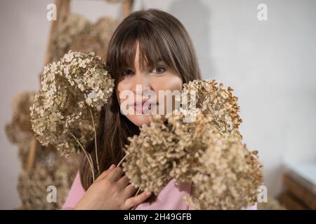Portrait of young dark-haired woman with bangs stands with flowers near window, hydrangea in her hands, profession - designer, florist. Stock Photo