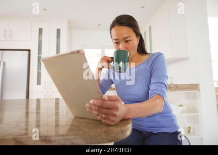 Focused asian woman drinking coffee and using tablet in kitchen Stock Photo