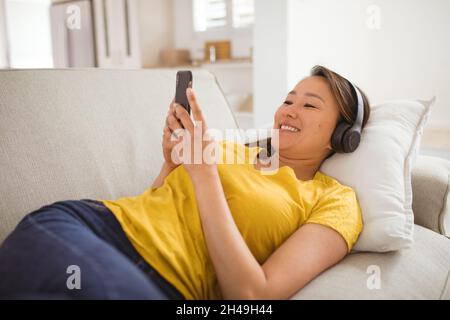 Happy asian woman wearing headphones lying on sofa with smartphone at home Stock Photo