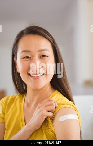 Portrait of happy asian woman sitting on sofa showing arm with plaster after vaccination Stock Photo