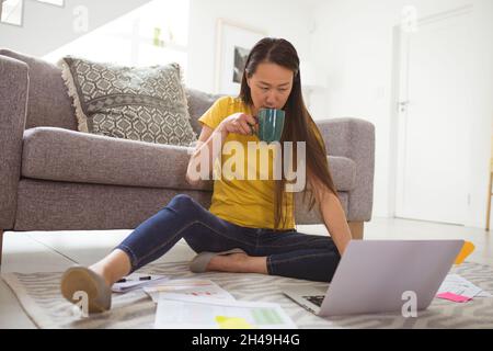 Focused asian woman drinking coffee and working remotely from home with smartphone and laptop Stock Photo