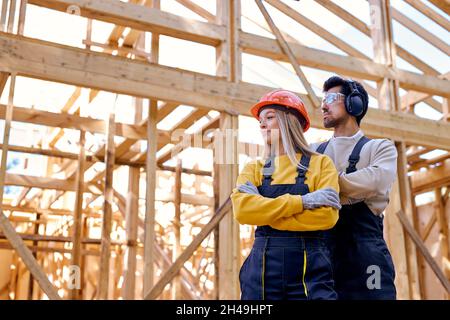 team of constructor engineers in New wooden residential construction home framing. Construction site. Beautiful caucasian lady and man in workwear, ha Stock Photo