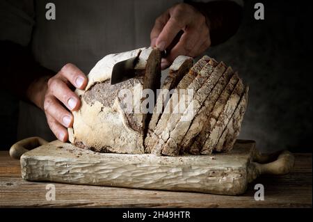 Male hands cut into slices a traditional loaf of bread on old wooden cutting board in dark setting, close-up. Stock Photo