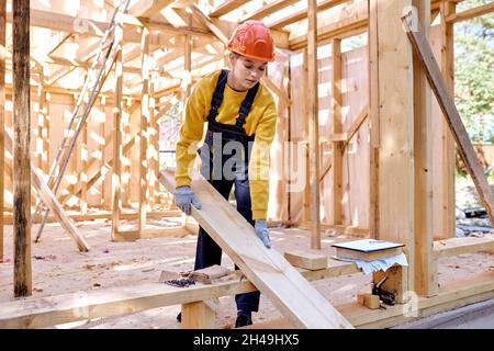 confident female constructor working on wooden construction site, wearing working uniform clothes, in gloves and hardhat, replacing wooden beam into a Stock Photo