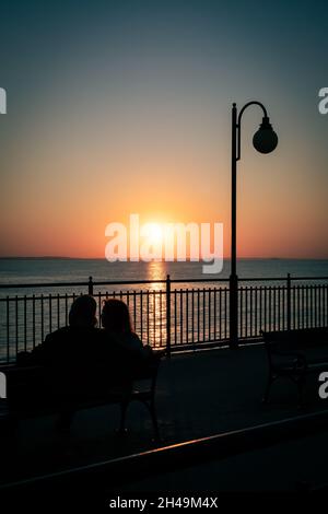 Miedzyzdroje, Poland - September 10, 2021. Silhouettes of a couple sitting on a bench on a pier, admiring sunset. Baltic sea, Miedzyzdroje, Poland. Stock Photo