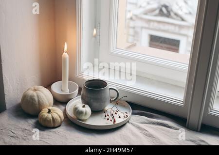 Autumn still life. Burning candle, white ceramic candleholder. Moody fall composition. White little pumpkins, cup of coffee and rose hips near window Stock Photo